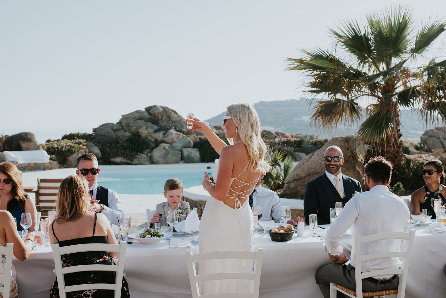 Mykonos wedding photographer: bride shot from behind raises a toast at the long reception table with guests smiling and palm tree in the background.