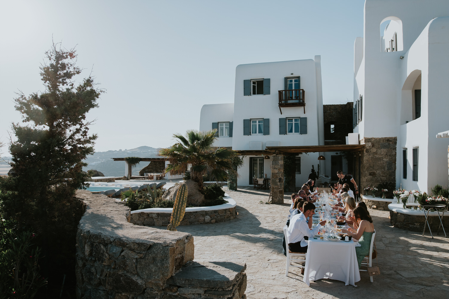 Mykonos wedding photographer: guests sat at the long reception table with villa in the background and a palm tree in afternoon light.