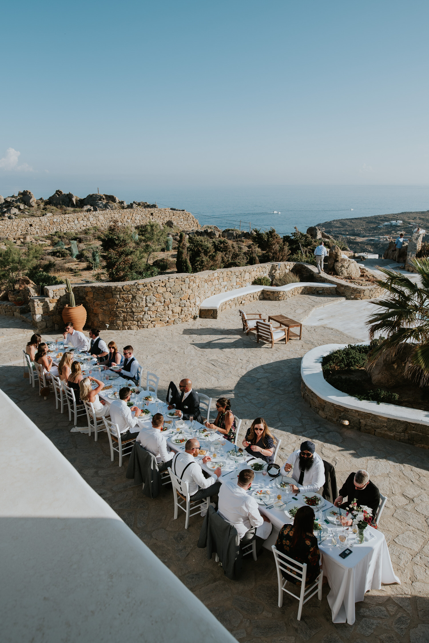 Mykonos wedding photographer: panoramic photo of guests sat at the long reception table with sea in the background.