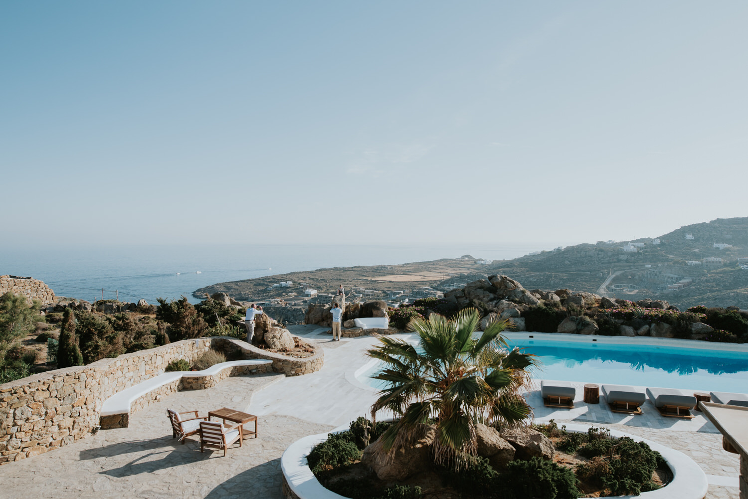 Mykonos wedding photographer: panoramic photo of the pool and a palm tree on reception terrace with sea in the background.