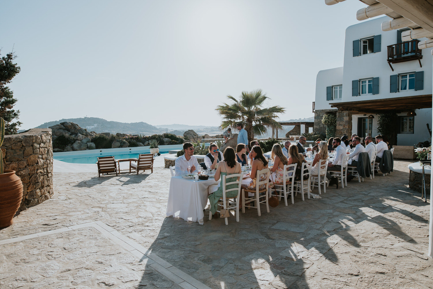Mykonos wedding photographer: reception terrace and the guests sat at the long table in front of the villa in the afternoon light with palm tree in the background.