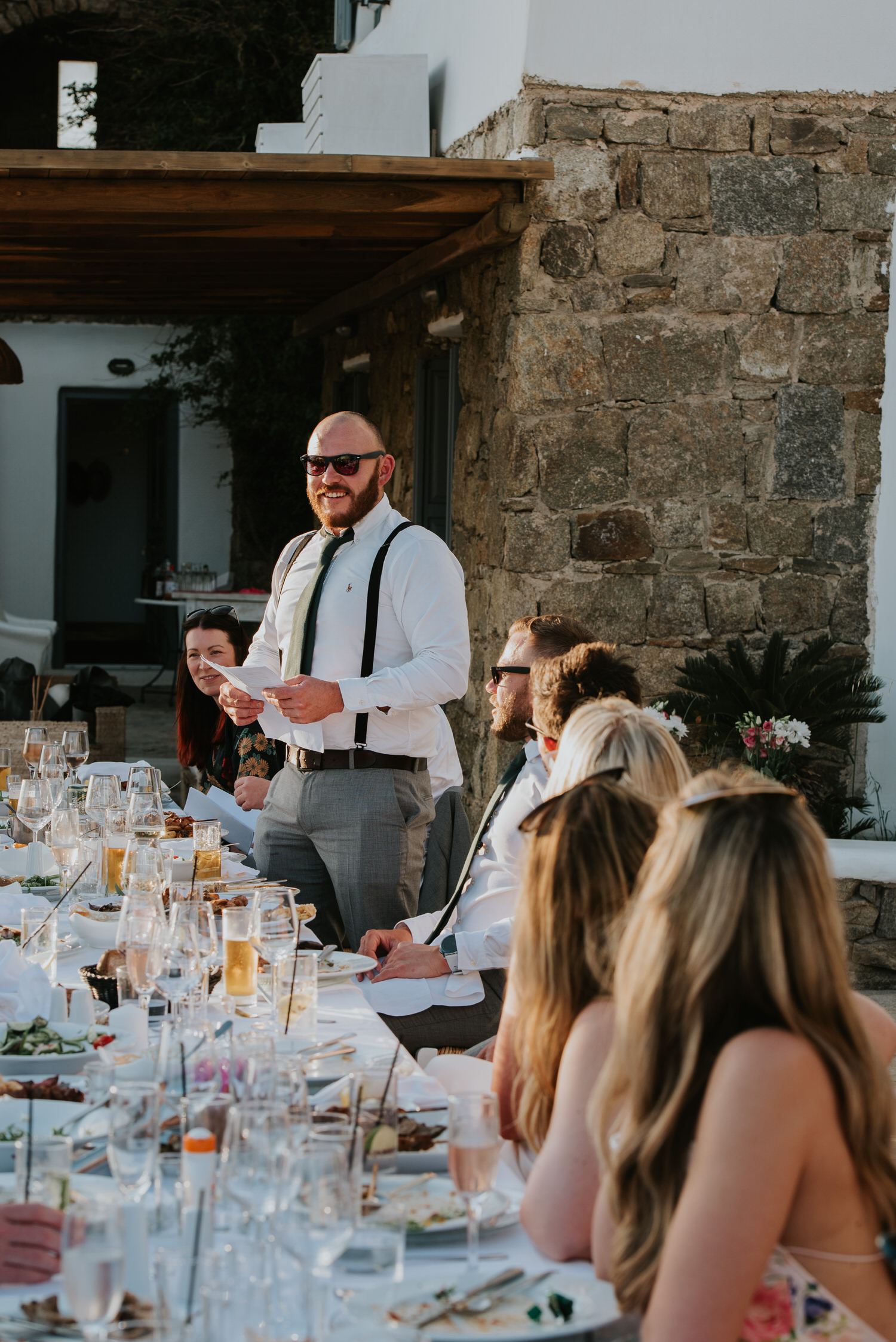 Mykonos wedding photographer: accent on best man in sunset light standing at the table during his speech.