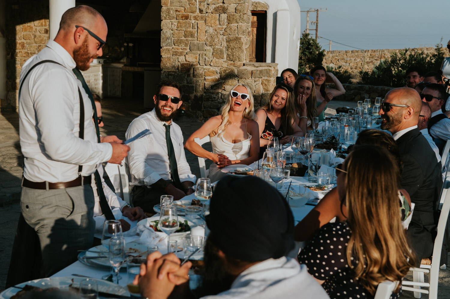 Mykonos wedding photographer: bride and groom and wedding guests sat at the table laughing listening to the best man's speech.