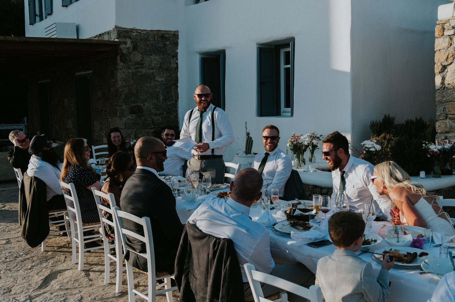 Mykonos wedding photographer: wedding guests sat at the table laughing listening to the best man's speech.