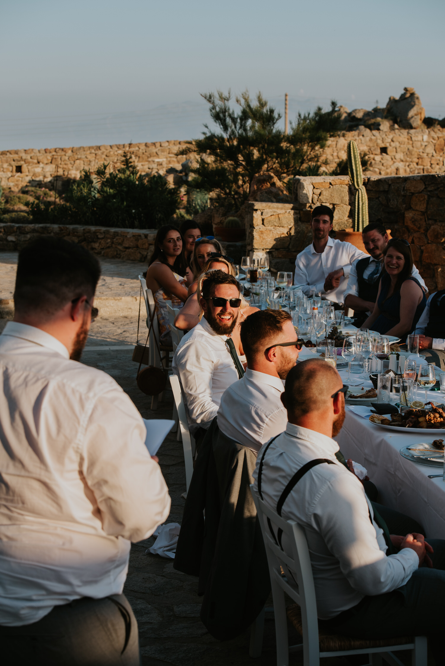 Mykonos wedding photographer: accent on the groom with wedding guests sat at the table laughing listening to the his best man's speech.
