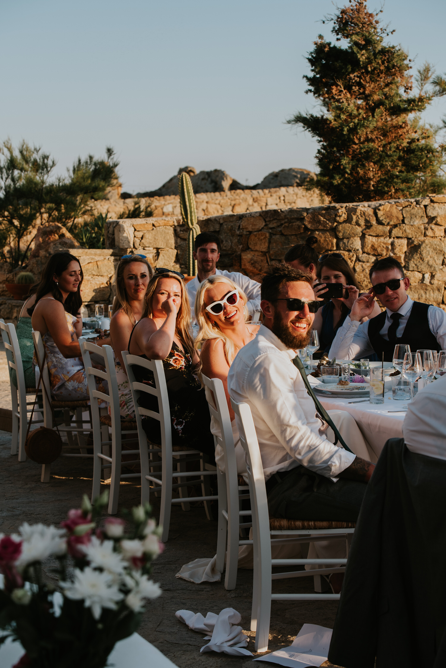 Mykonos wedding photographer: bride and groom with wedding guests sat at the table in sunset light listening to the speeches.