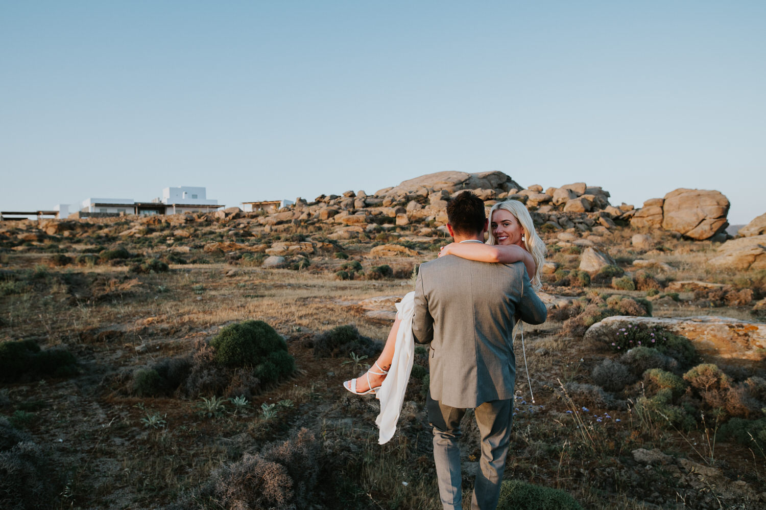 Mykonos wedding photographer: groom holding his bride in his arms walking through grassy field towards the massive rocks in sunset light.