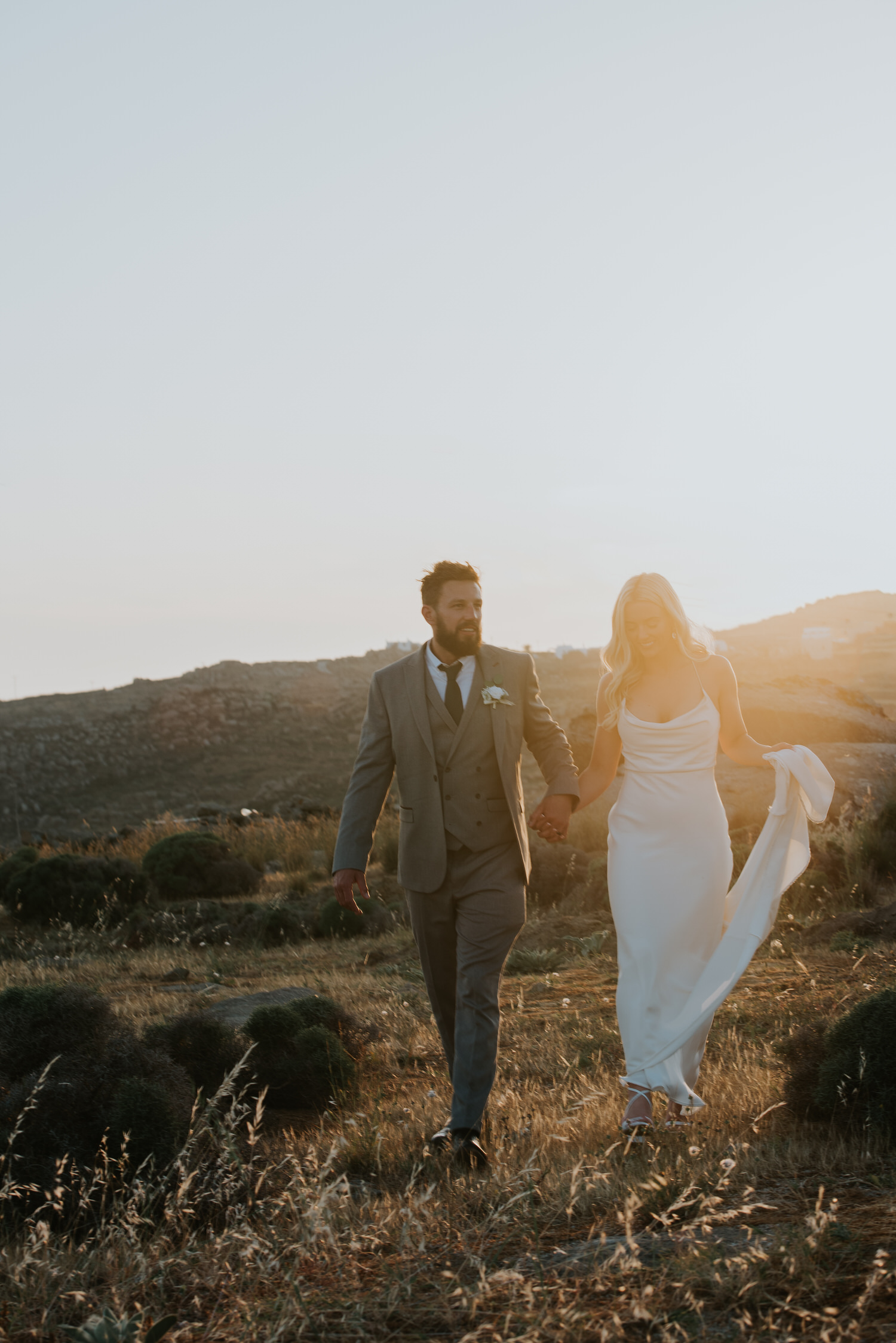 Mykonos wedding photographer: bride and groom basked in beautiful golden light walking through grassy field.