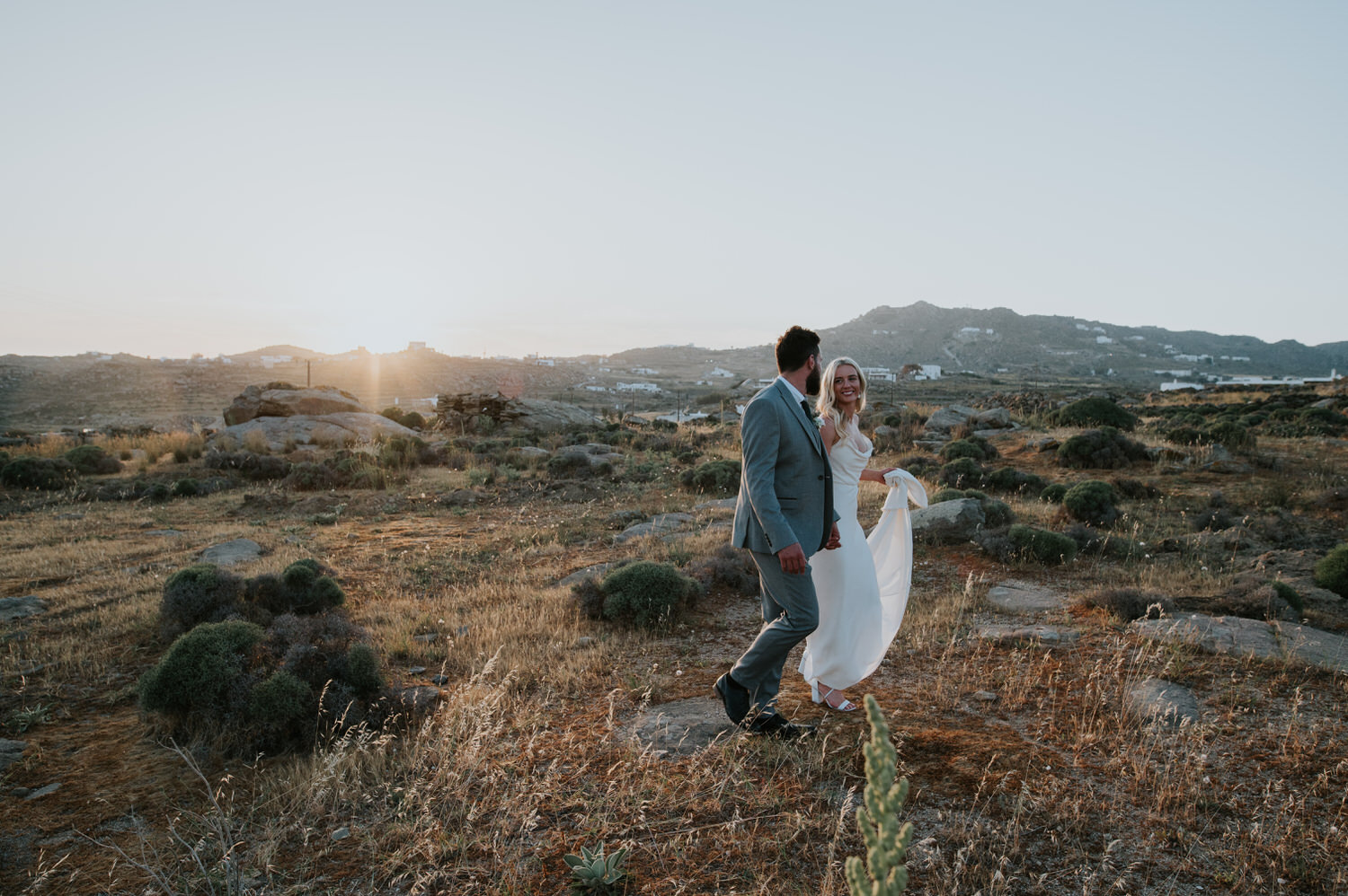 Mykonos wedding photographer: bride and groom looking at each other walking in sunset light through grassy field.