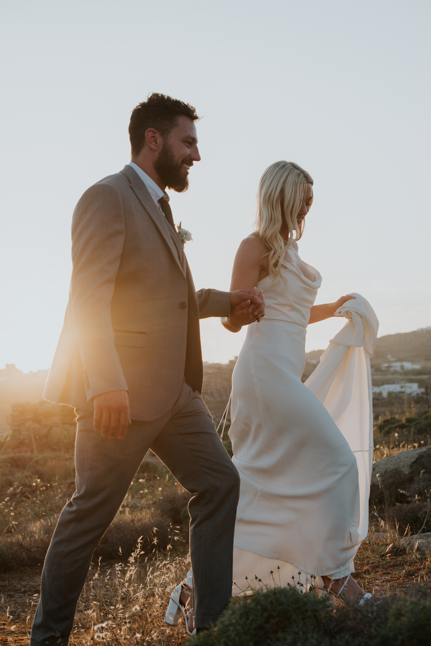Mykonos wedding photographer: bride and groom walking through grassy field in beautiful golden light.
