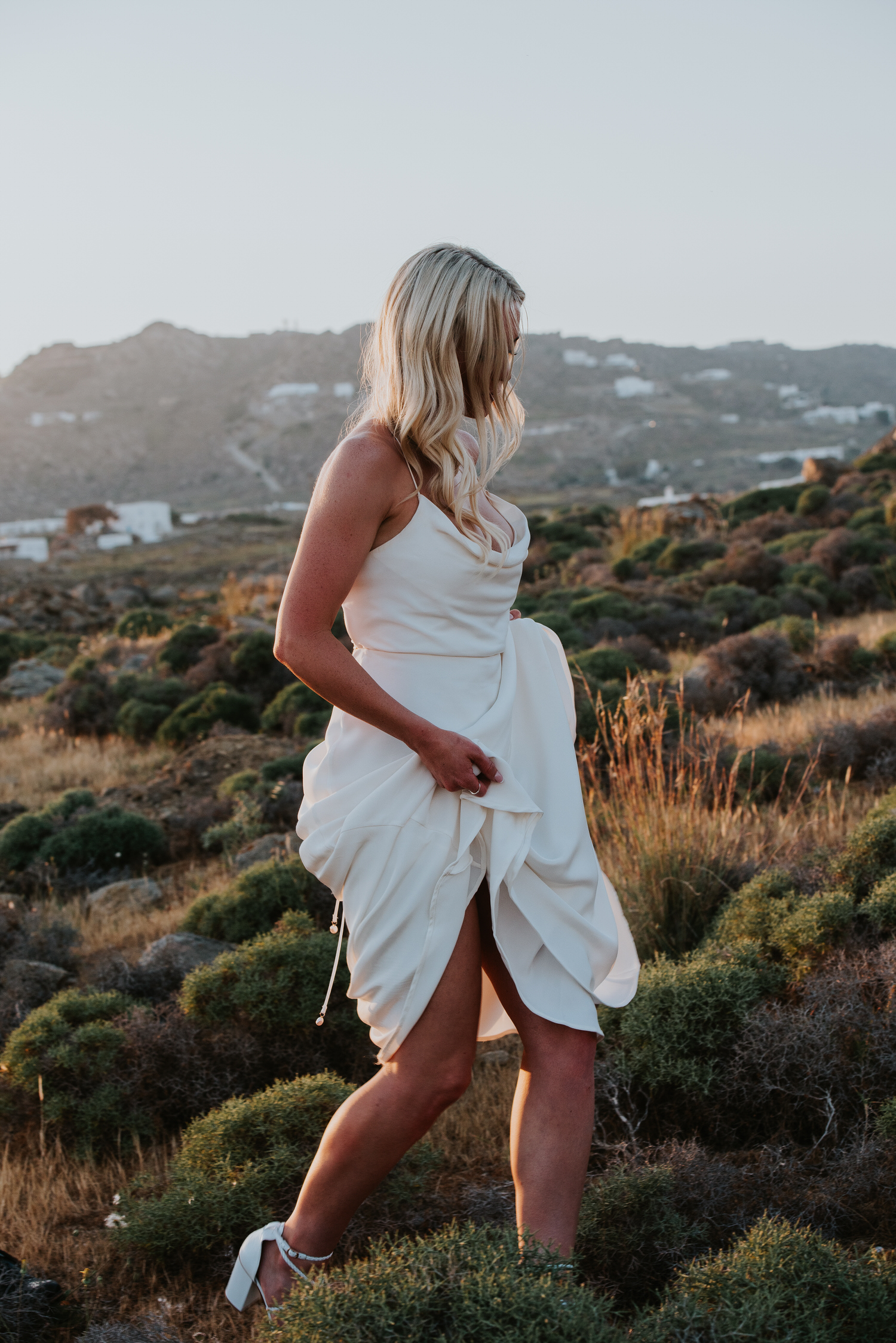 Mykonos wedding photographer: bride walking in sunset light with her dress up in her hands walking through grassy field.