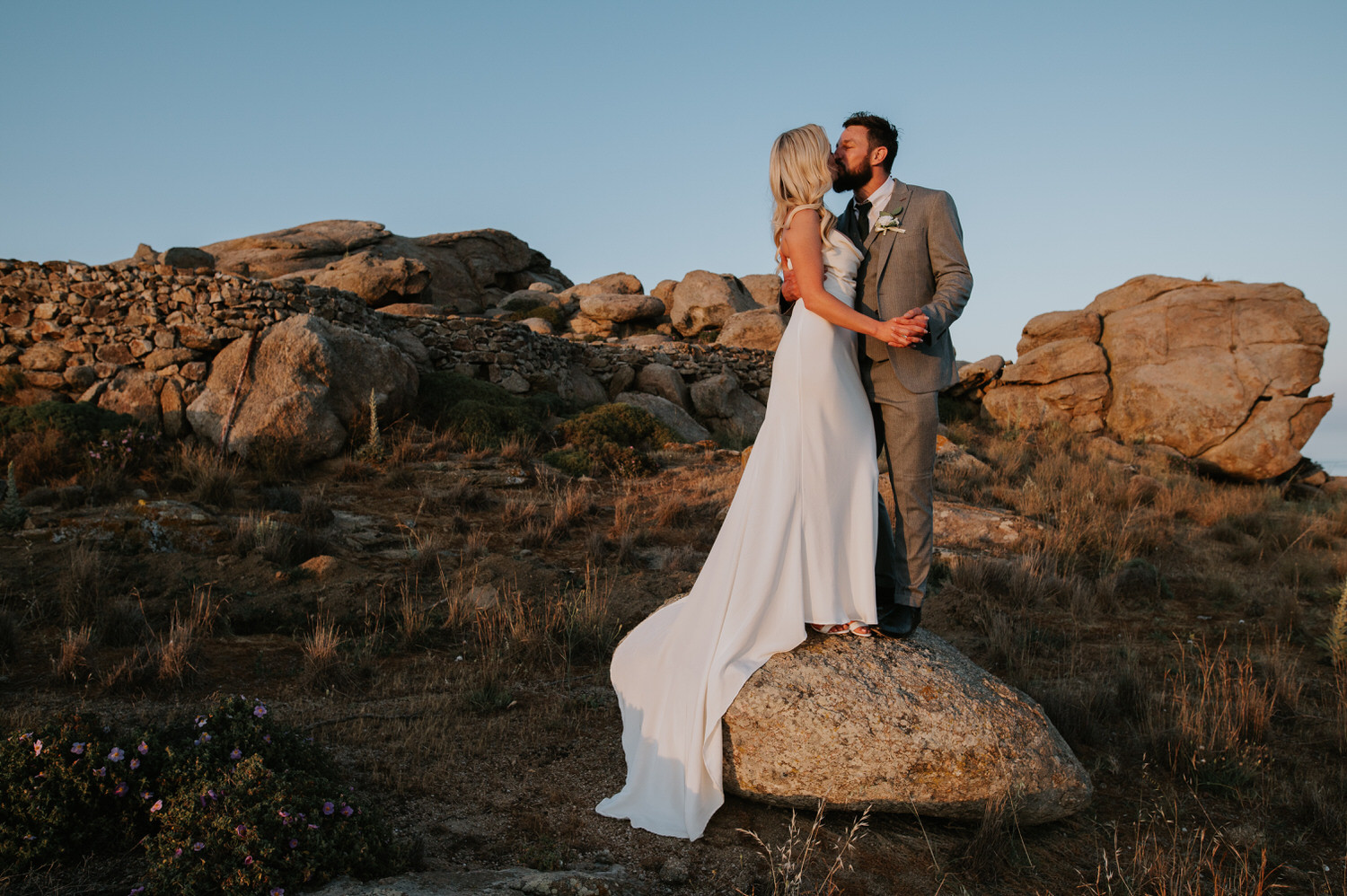 Mykonos wedding photographer: bride and groom kissing holding hands standing on the rock surrounded by the landscape.