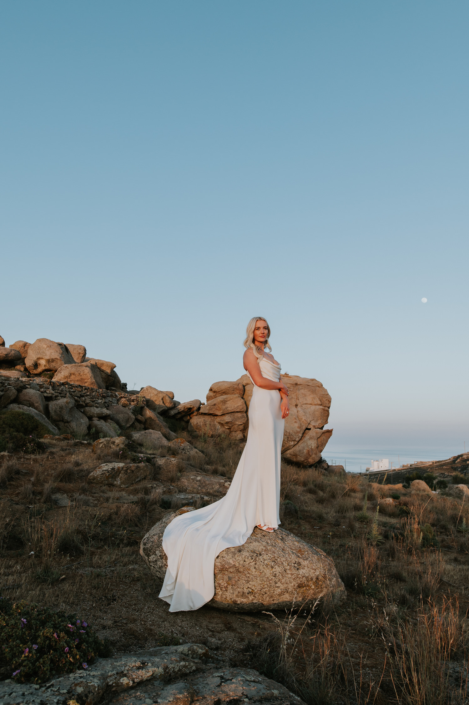 Mykonos wedding photographer: bride standing in her beautiful dress on the rock surrounded by the landscape with the moon in the background.
