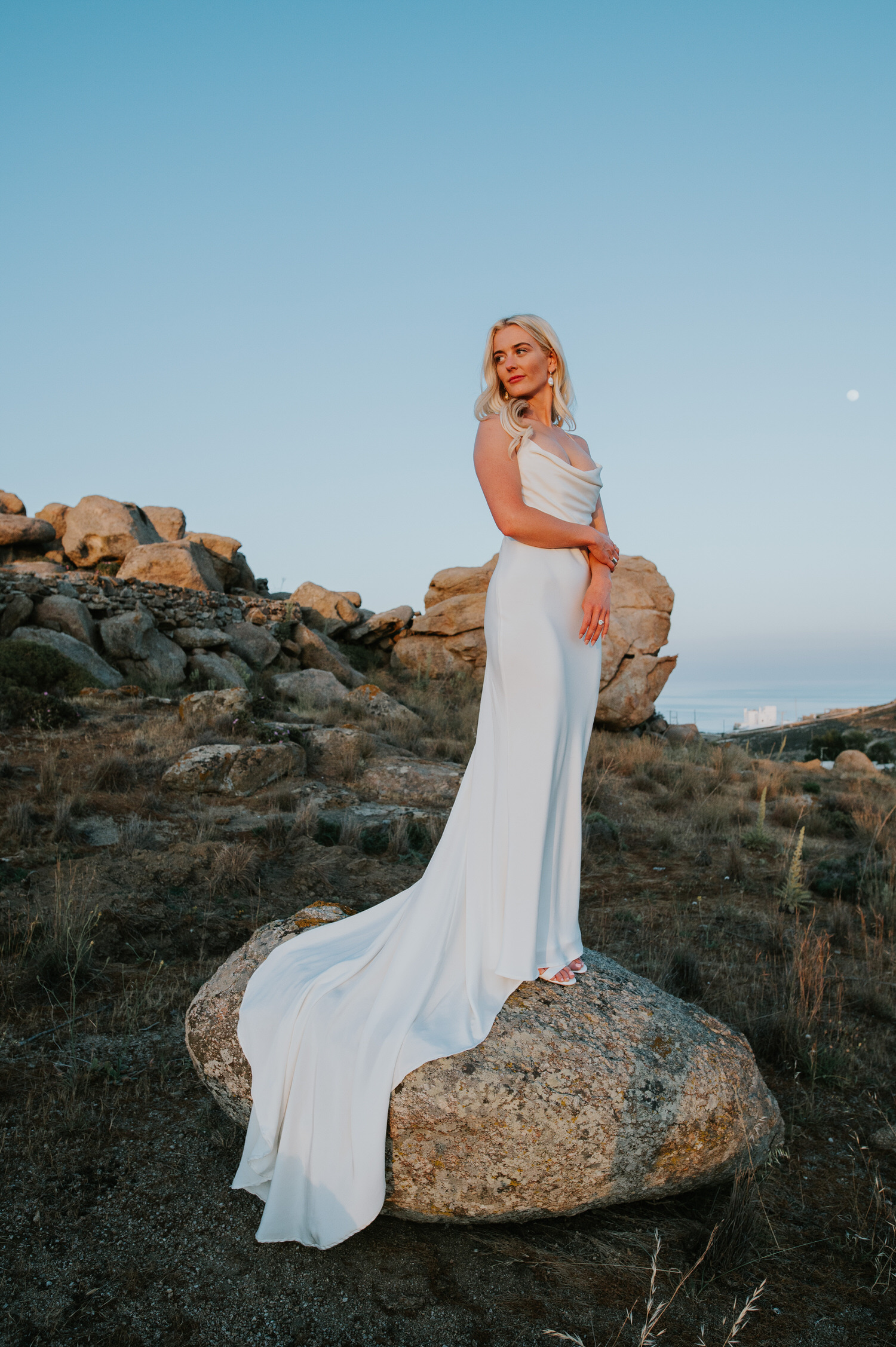 Mykonos wedding photographer: bride standing in her beautiful dress on the rock looking away in the landscape with the moon in the background.
