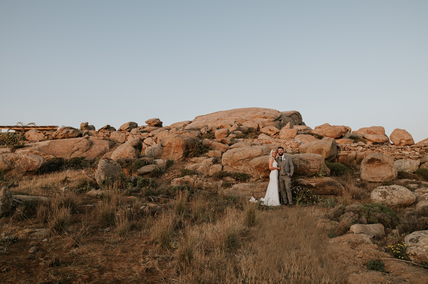 Mykonos wedding photographer: bride and groom standing in the grass field next to the giant rocks after the sunset.