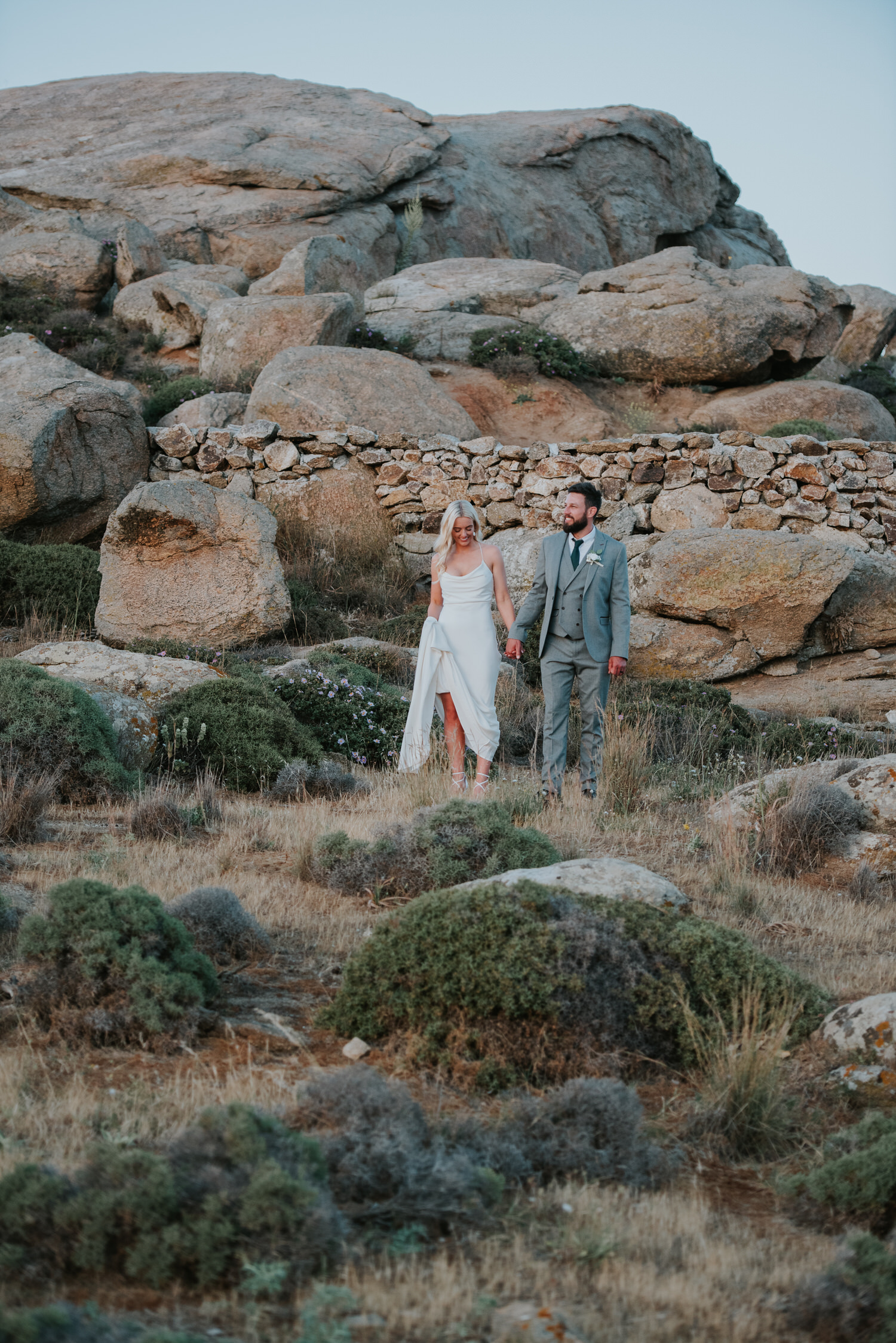 Mykonos wedding photographer: bride and groom in Mykonian landscape surrounded by rocks walking through grass.