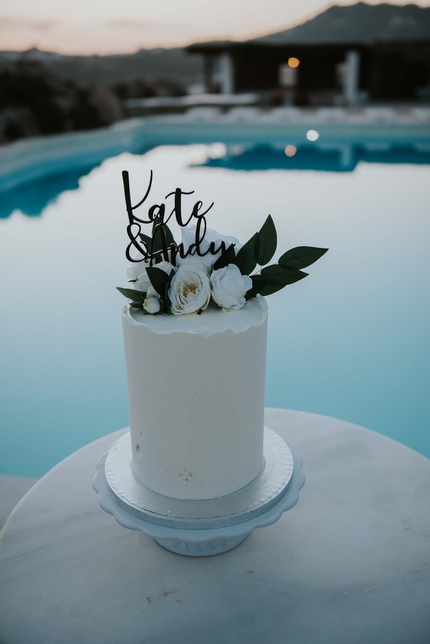 Mykonos wedding photographer: close up of wedding cake with bride and groom's fingerprints next to turquoise swimming pool on their Mykonos wedding.