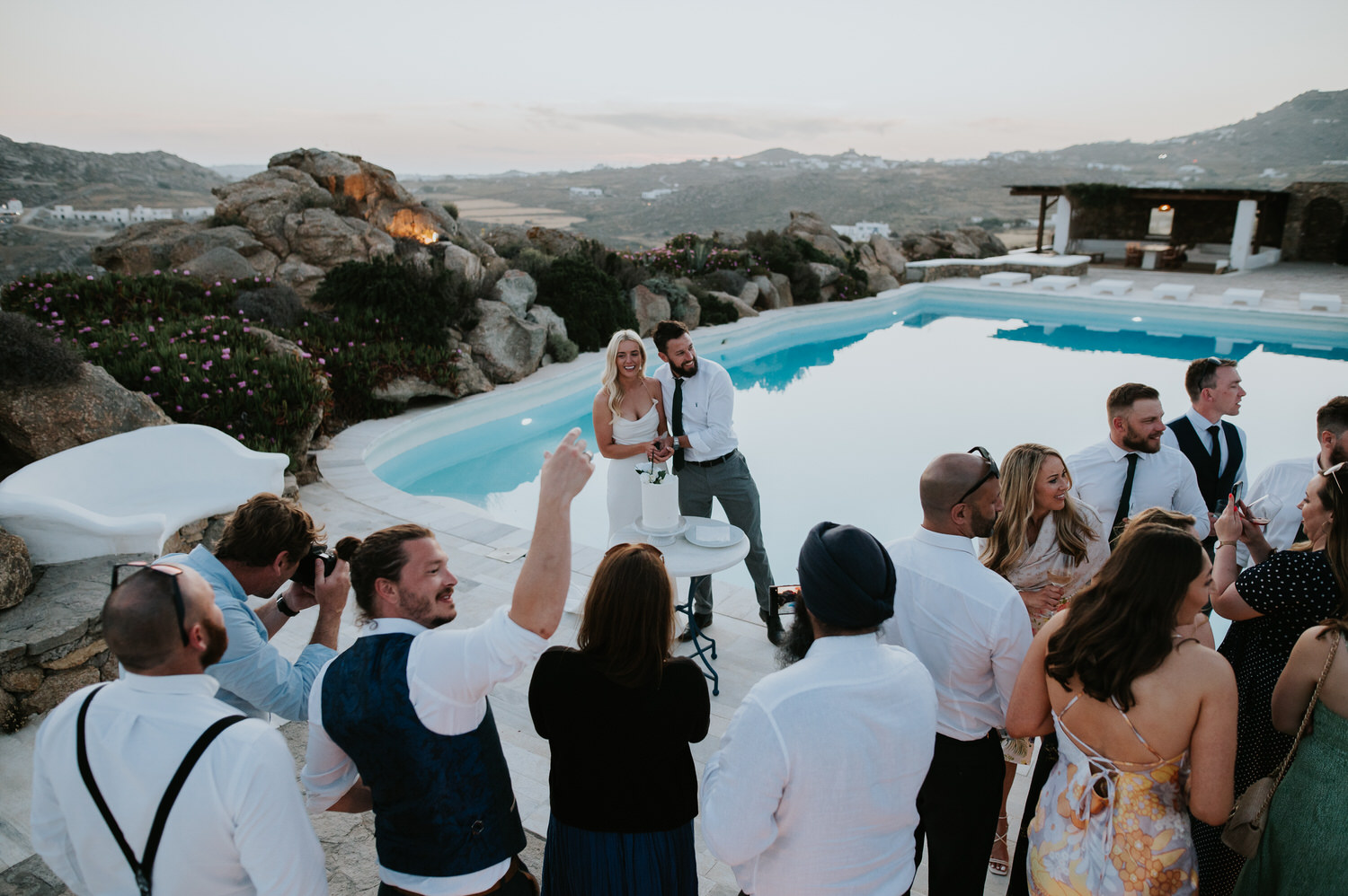 Mykonos wedding photographer: bride and groom standing with everyone and a cake next to turquoise swimming pool on their Mykonos wedding.