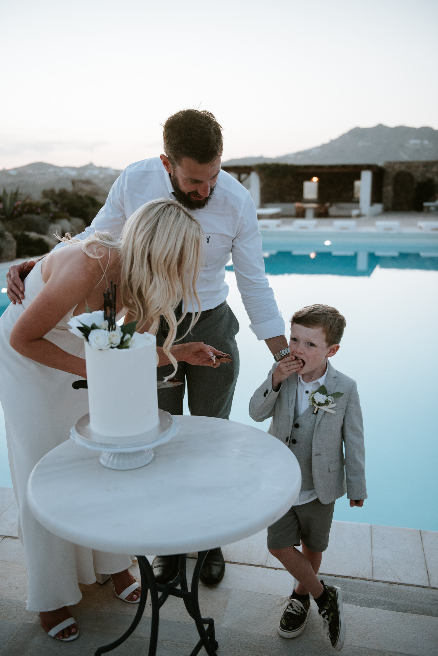 Mykonos wedding photographer: kid testing the cake standing next to bride and groom with pool in the background.