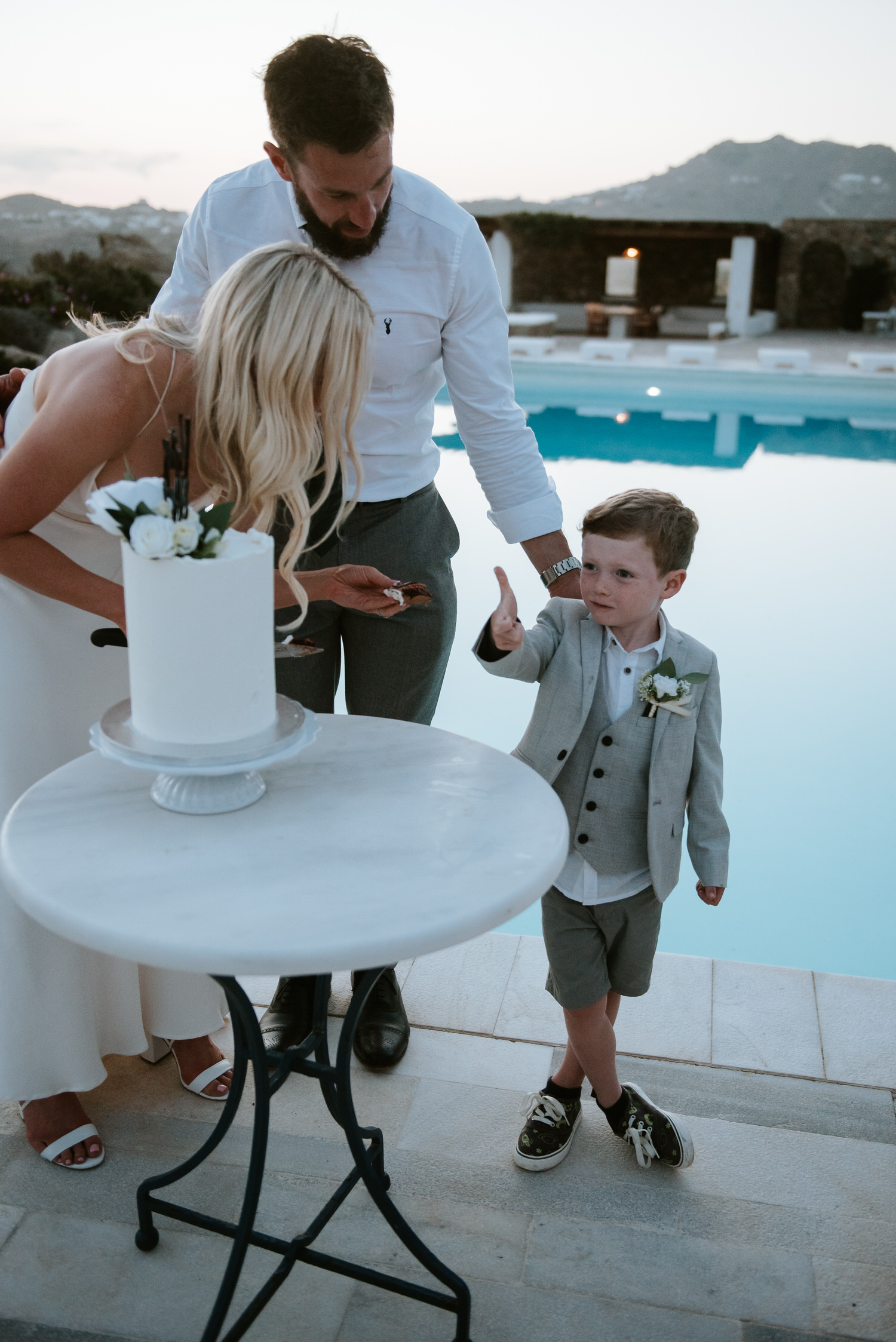 Mykonos wedding photographer: kid gives thumbs up for the cake standing next to bride and groom with pool in the background.
