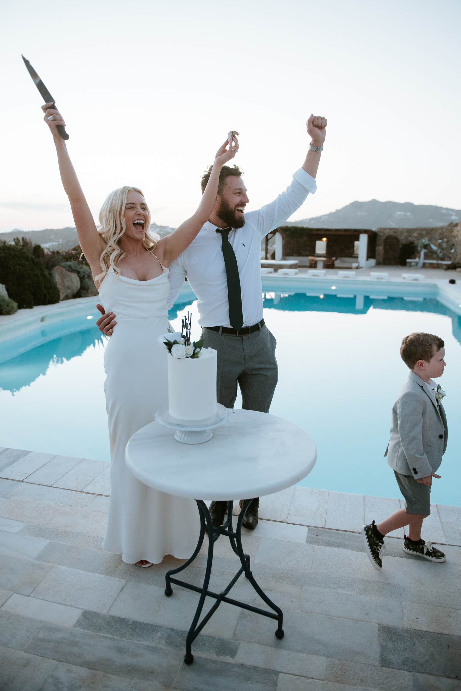 Mykonos wedding photographer: bride and groom screaming with delight with hands in the air as kid walks away from the cake.