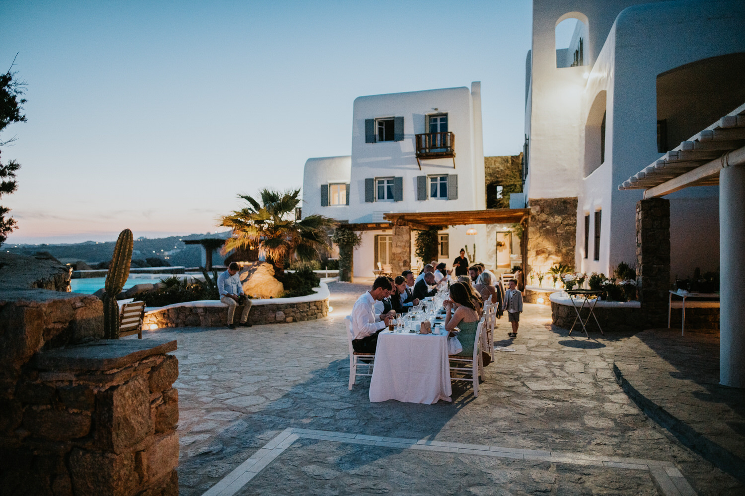 Mykonos wedding photographer: wedding party at the table with the villa, palm tree and the turquoise swimming pool at dusk on Mykonos wedding.