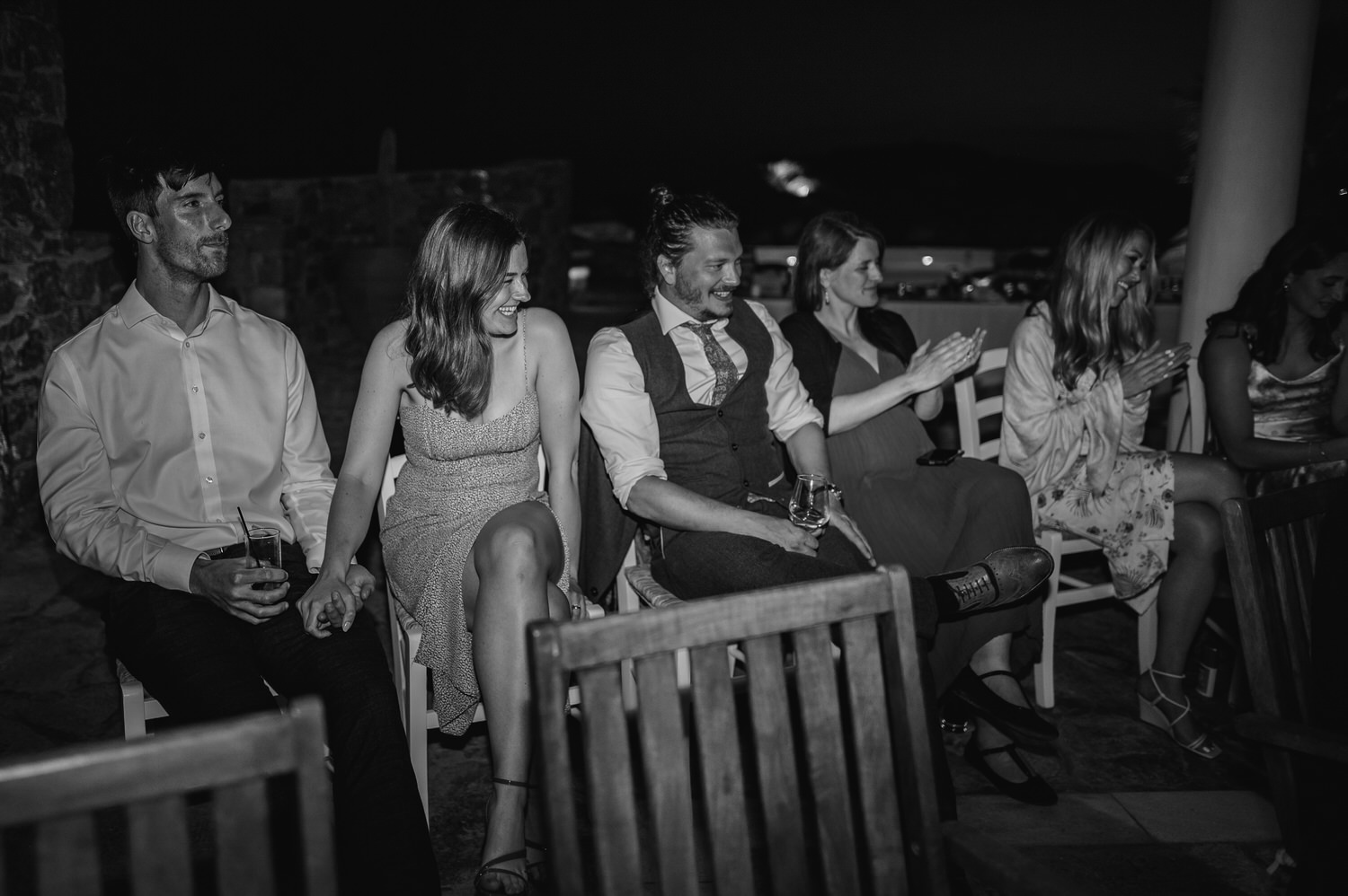 Mykonos wedding photographer: black and white photo of guests laughing sat around the groom during the speeches on Mykonos wedding.
