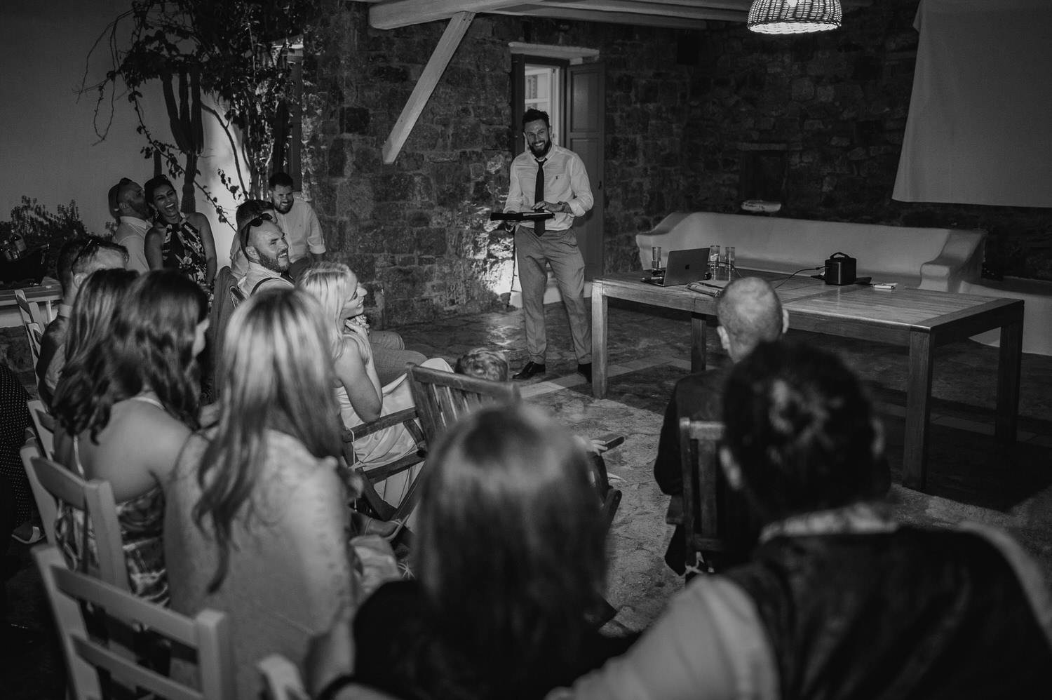 Mykonos wedding photographer: black and white photo of groom surrounded by guests during the speeches on Mykonos wedding.