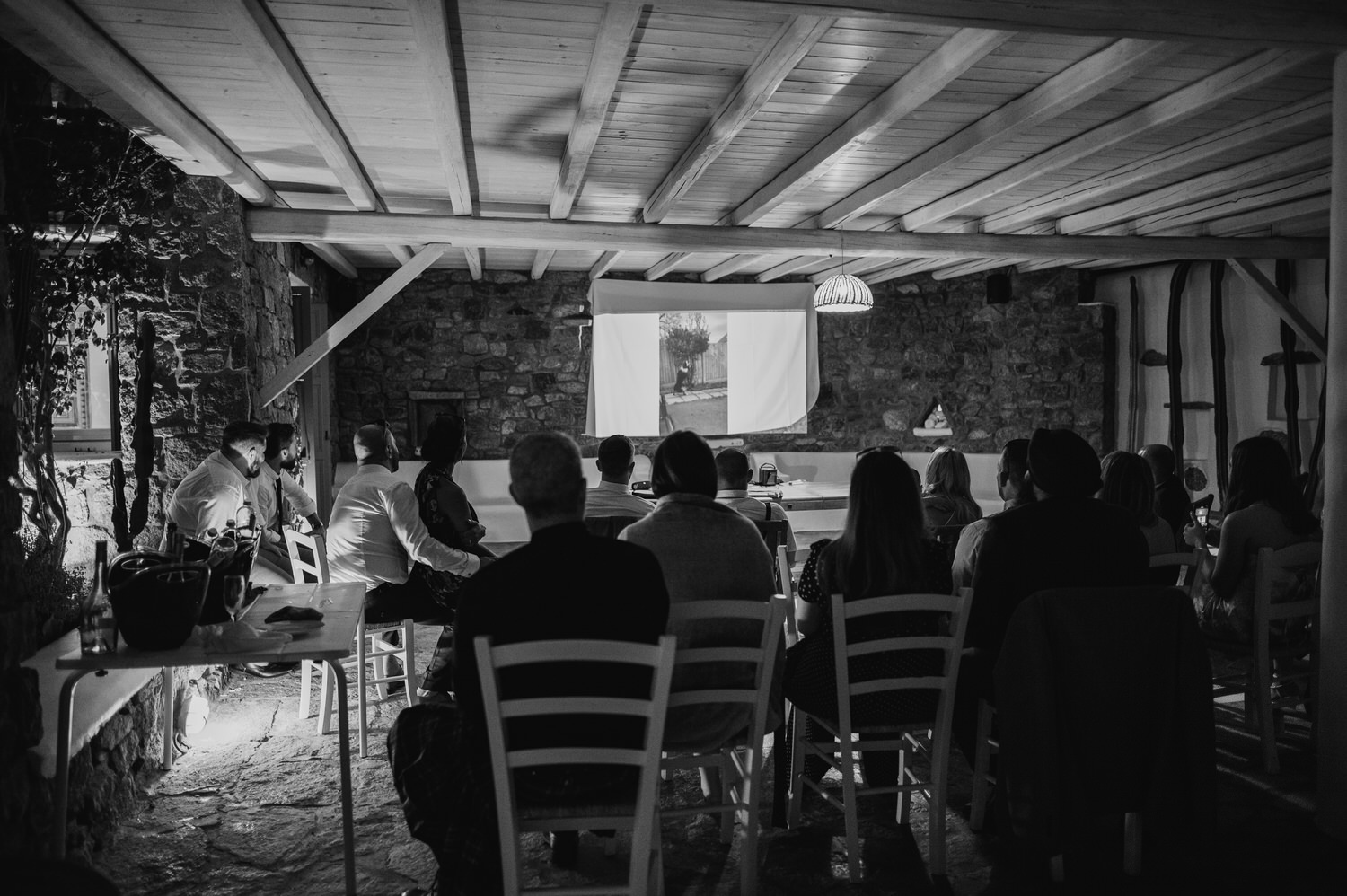 Mykonos wedding photographer: black and white photo of the whole wedding party watching pre recorded wedding wishes.