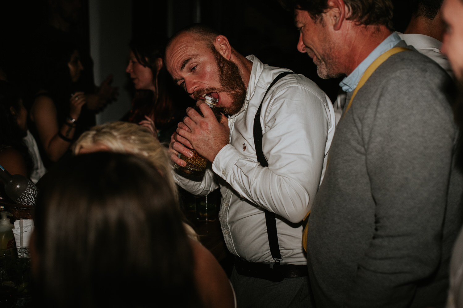 Mykonos wedding photographer: wedding guest skilfully opening a bottle with his teeth on dance floor for Mykonos wedding celebration.