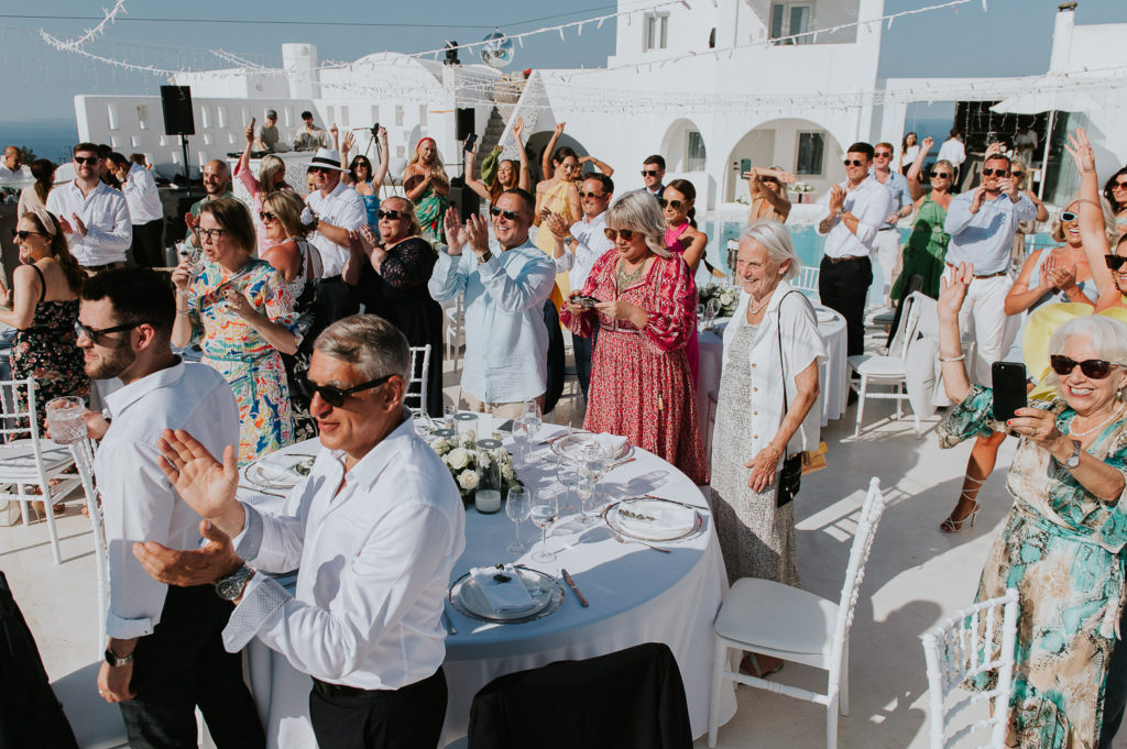 Rocabella Santorini Wedding: guests standing up cheering and clapping as the couple enters the reception by Wedding photographer Santorini.