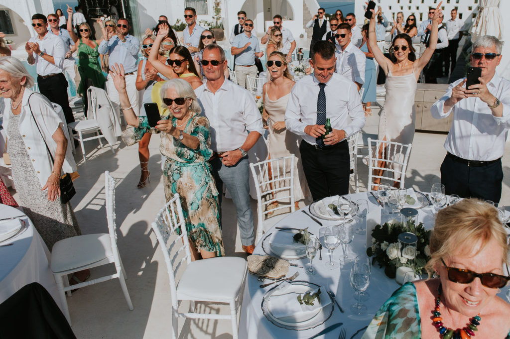 Rocabella Santorini Wedding: guests standing up cheering as the couple enters the reception by Wedding photographer Santorini.