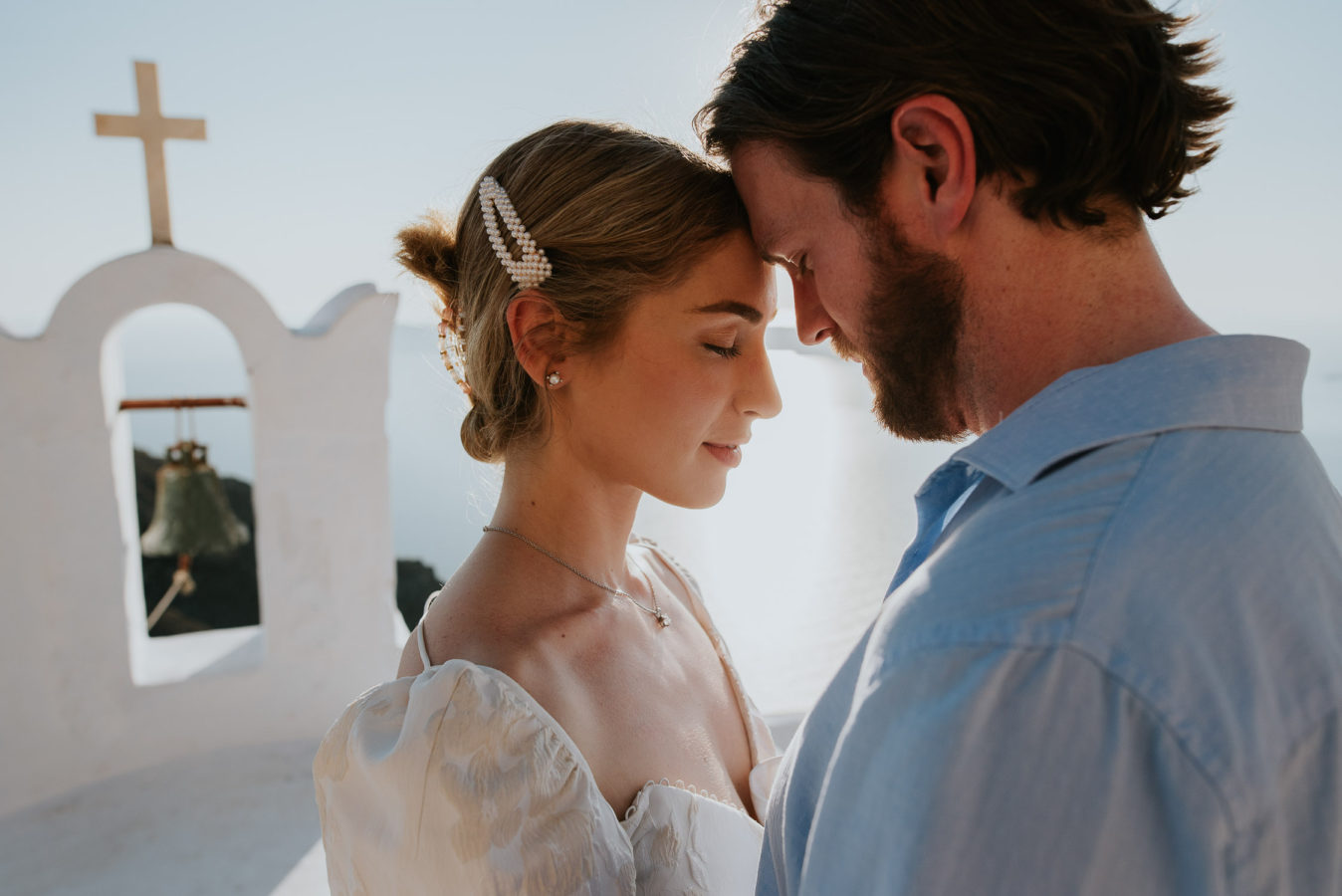 Wedding photographer Santorini: closeup of a couple gently touching their foreheads bathed in the sun with bell tower in the background by Ben and Vesna.