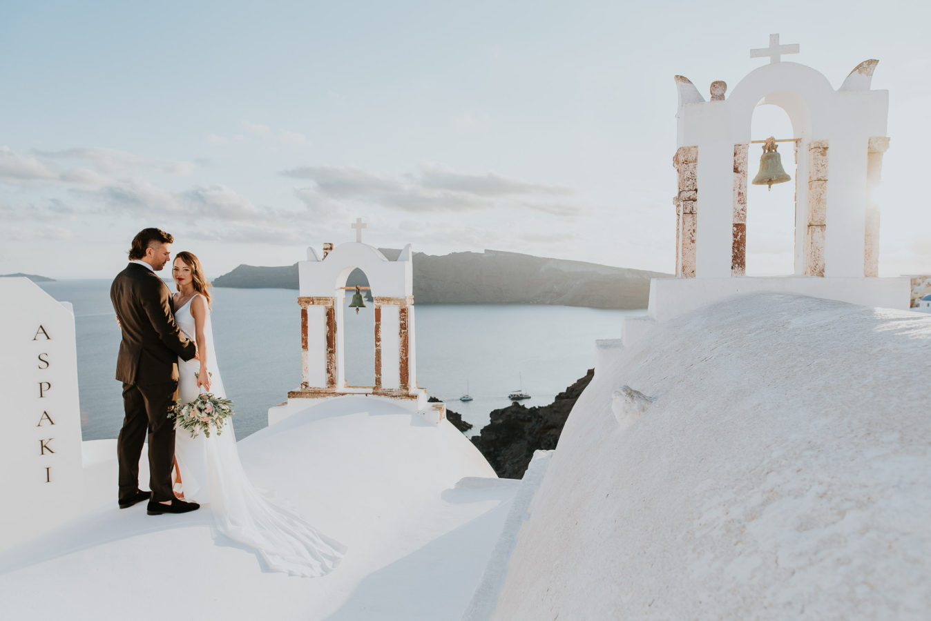 Wedding photographer Santorini: panoramic photo of bride and groom on the church rooftop with the bell towers in the background by Ben and Vesna.