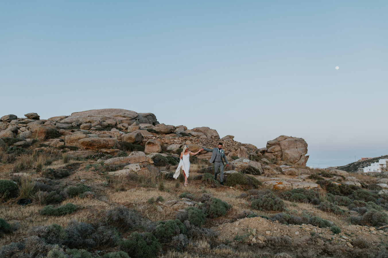 Wedding photographer Santorini: bride and groom in the filed walking holding hands at dusk surrounded by rocks and wild bushes by Ben and Vesna.