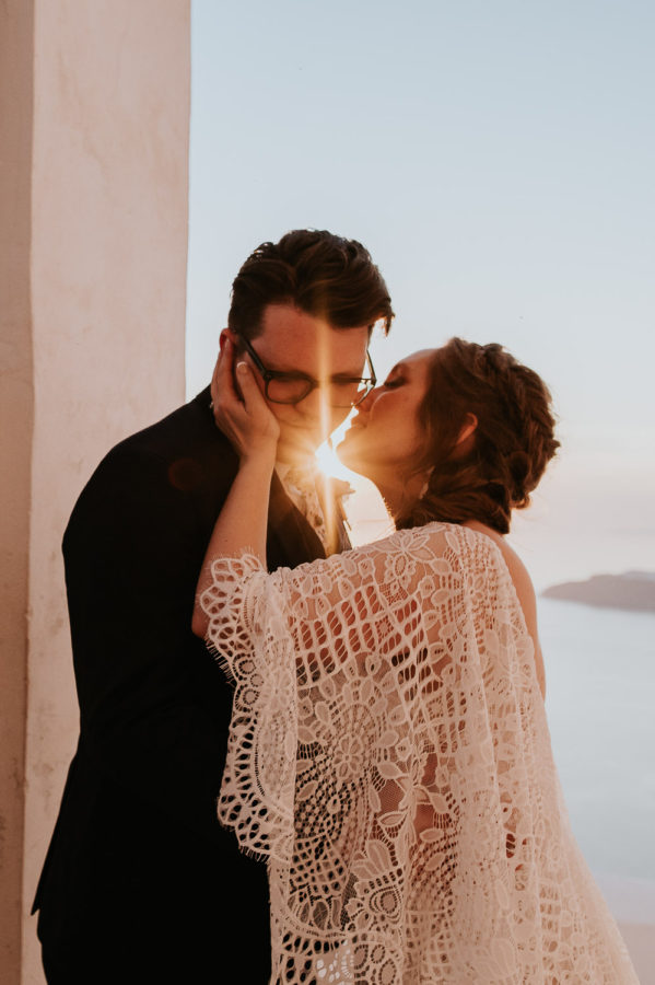 Wedding photographer Santorini: bride in a boho dress reaches to kiss the groom's cheek with sun flare between them by Ben and Vesna.